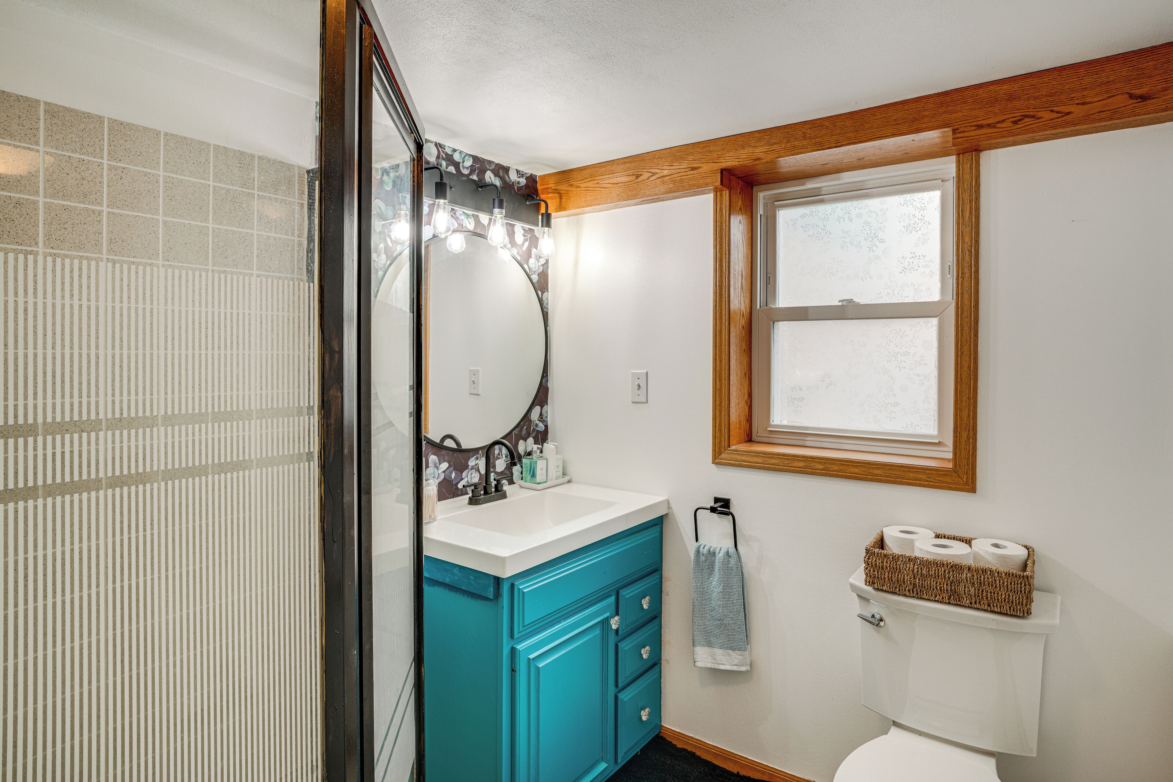 Une salle de bain avec des toilettes blanches, un meuble-lavabo bleu avec un miroir et une fenêtre située au-dessus des toilettes.
