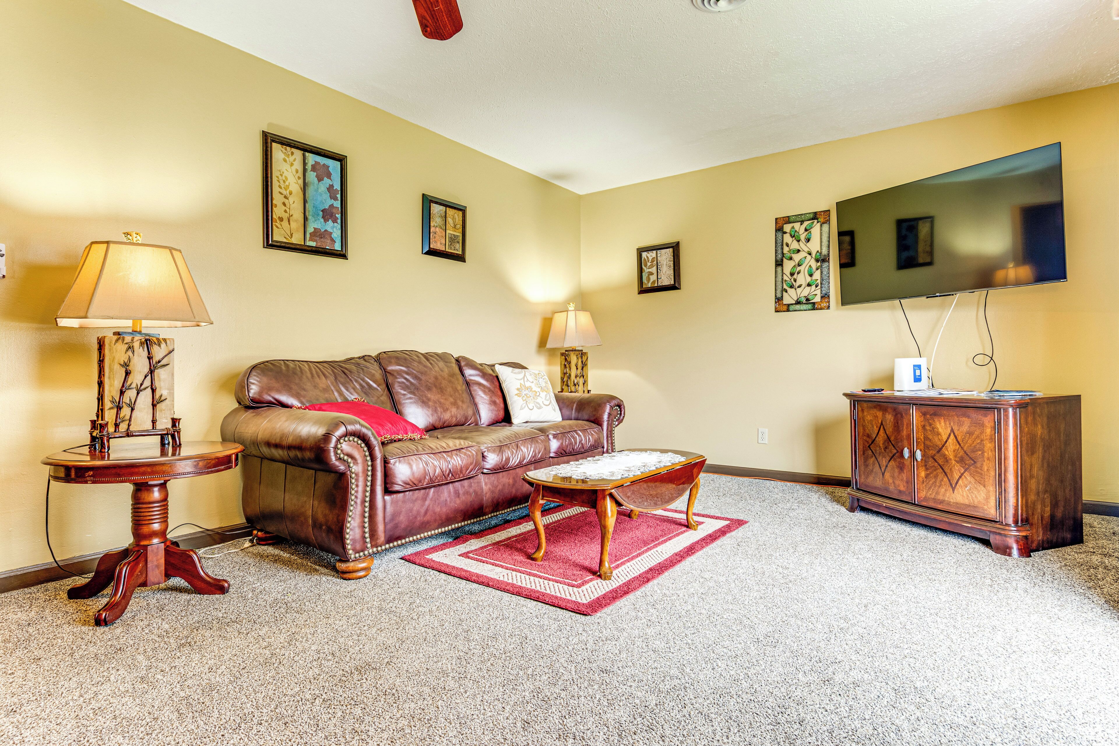 A living room with a leather sofa, a wooden coffee table, a red rug, and a flat-screen TV mounted on the wall.