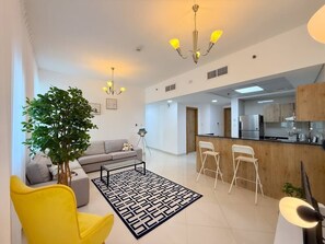 A modern living room with a sofa, yellow armchair, dining area, kitchen, and a large tree. - Beach Lux New 2BD | Marina view | CEG Group (Dubai)