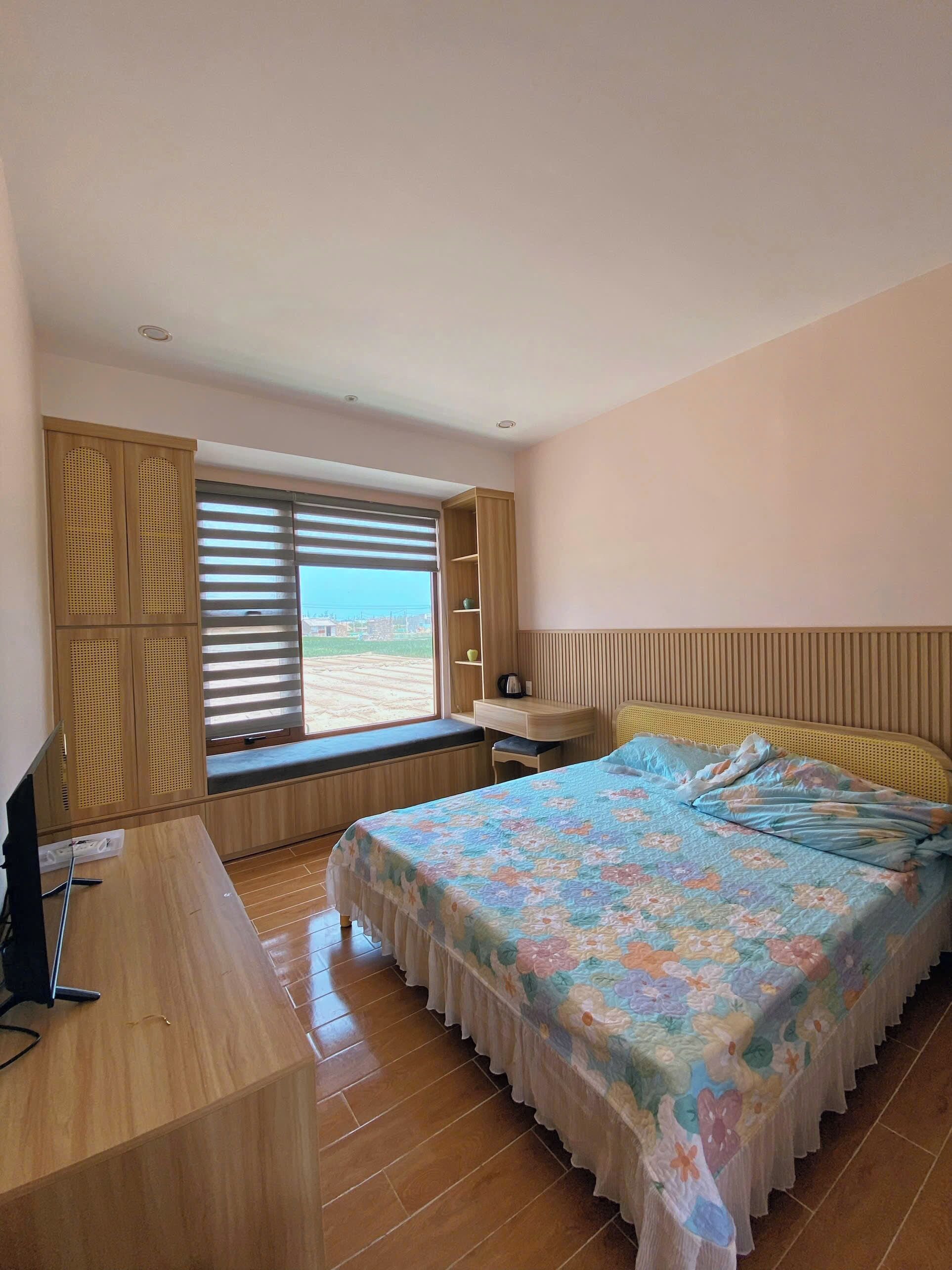A bedroom with a bed, a desk, a TV, a window with blinds, and a view of the outside.