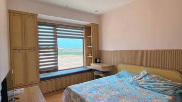 A bedroom with a bed, a desk, a TV, a window with blinds, and a view of the outside.