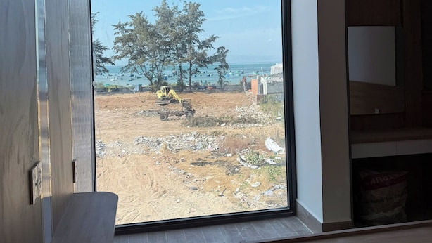 A room with a large window, a wooden floor, and a view of a construction site with a tree and the sea.