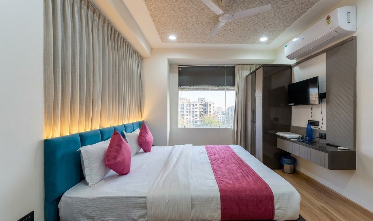 A hotel room with a bed, curtains, a television, an air conditioning unit, and a window with a city view.