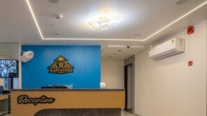 A reception desk with a blue wall and a logo, a flat-screen TV, and an air conditioning unit on the wall. - Hotel Rajvanshi Inn (Ahmedabad)
