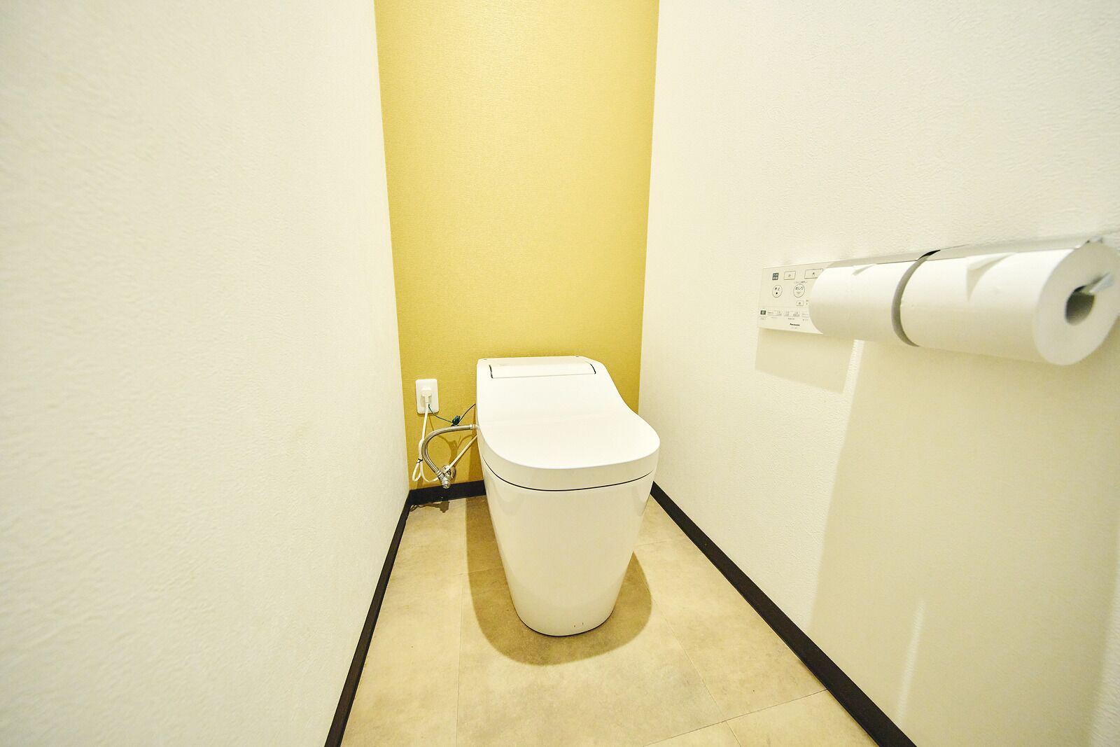 Un baño pequeño y estrecho con inodoro, porta papel higiénico y un enchufe visible.
