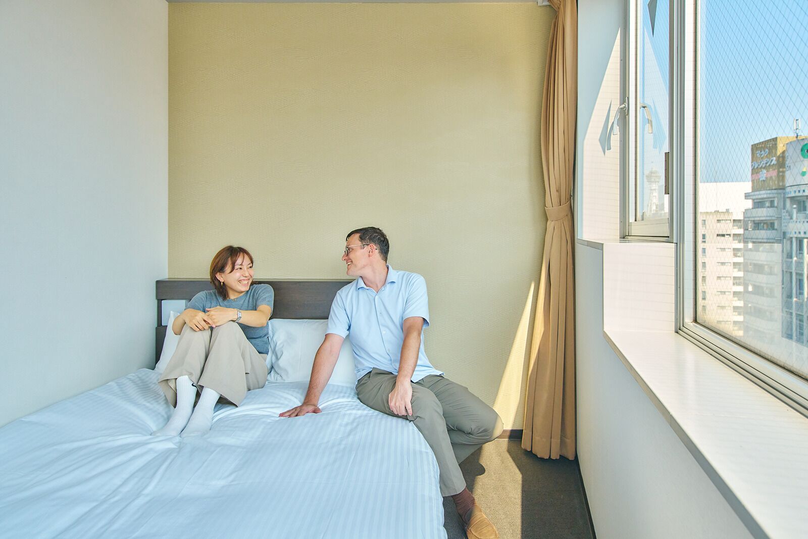 A hotel room with a bed, a window with curtains, and a view of buildings outside.