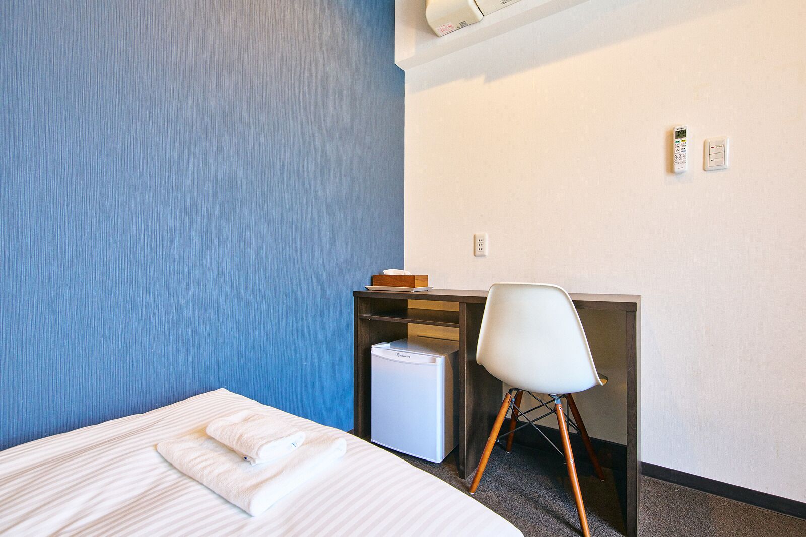 A hotel room with a bed, a desk, a chair, and a small white trash bin.