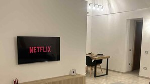 A modern living room with a flat-screen TV mounted on a white wall, a wooden bench, a grey sofa, and a dining area with a table and chairs. - Domus Gazzara, cozy and comfortable vacation home in the heart of the historic center (Napoli)