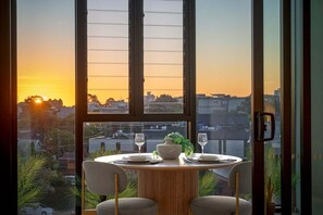 Ein Essbereich mit Tisch für zwei Personen, einer Vase und Blick auf den Sonnenuntergang durch große Fenster.