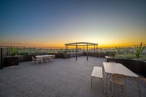 Eine Terrassensitzgelegenheit mit Tischen und Stühlen, einer Pergola und Blick auf den Sonnenuntergang über einer Stadtlandschaft.