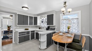 A modern kitchen with grey cabinets, a wooden dining table, and a window with floral curtains. - Irish Safari Retreat / Walk to Zoo / Minutes to Notre Dame (South Bend)