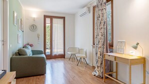 Ein Zimmer mit einer Couch, einem Schreibtisch mit Lampe, einem Stuhl, einer Uhr und einer Tür, die nach draußen führt.