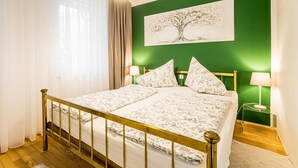 Un lit recouvert d’une couvre-lit à motifs floraux, un mur vert orné d’un tableau représentant un arbre, et deux lampes de chevet.