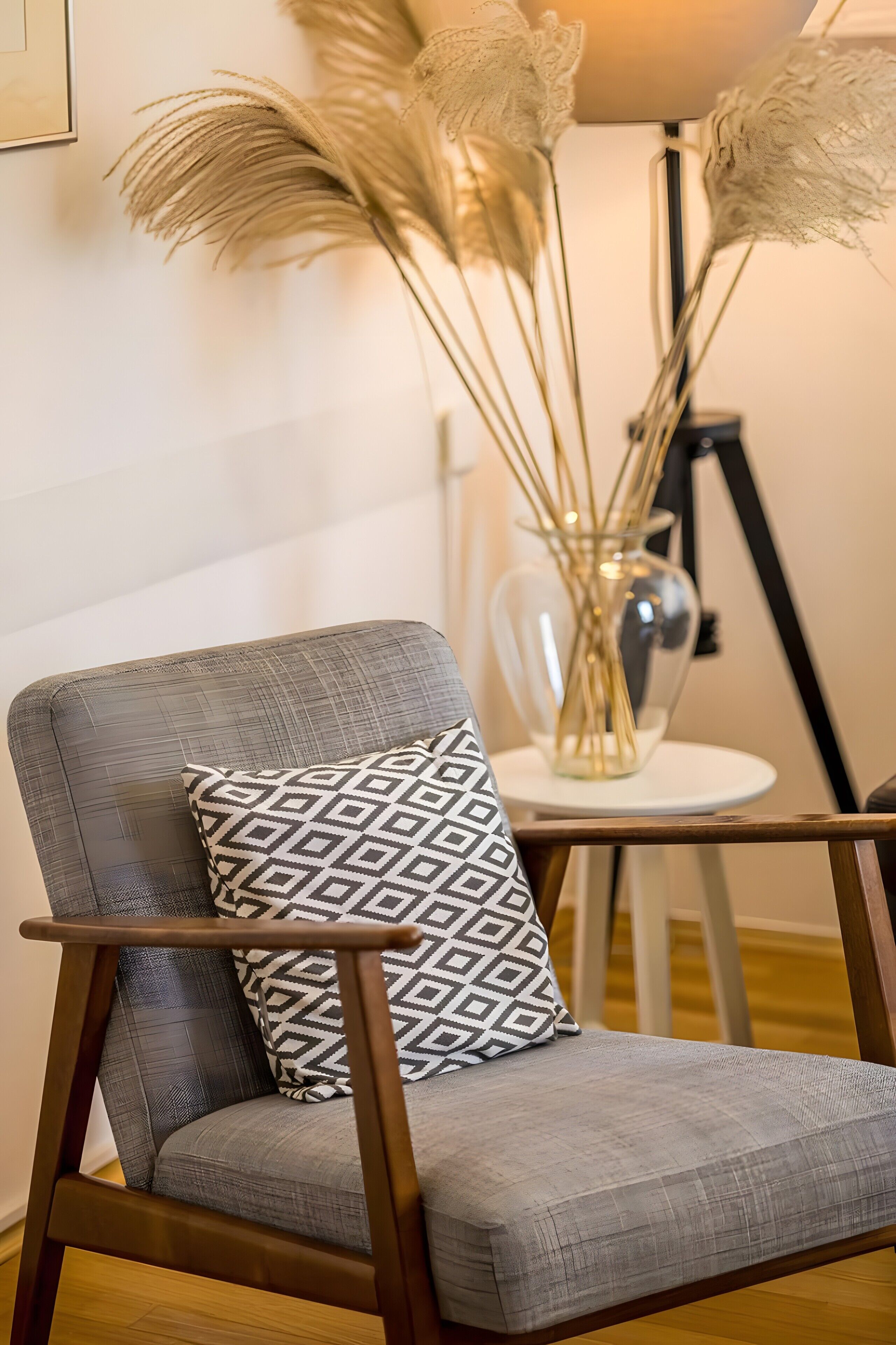 Una silla moderna con un cojín geométrico, un florero con plantas altas y una lámpara de pie en una habitación con suelo de madera.