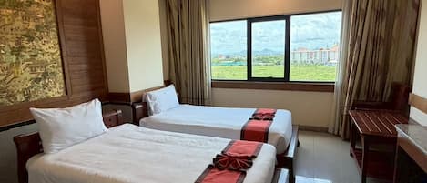 A hotel room with two beds, a desk, and a large window offering a view of buildings and greenery.