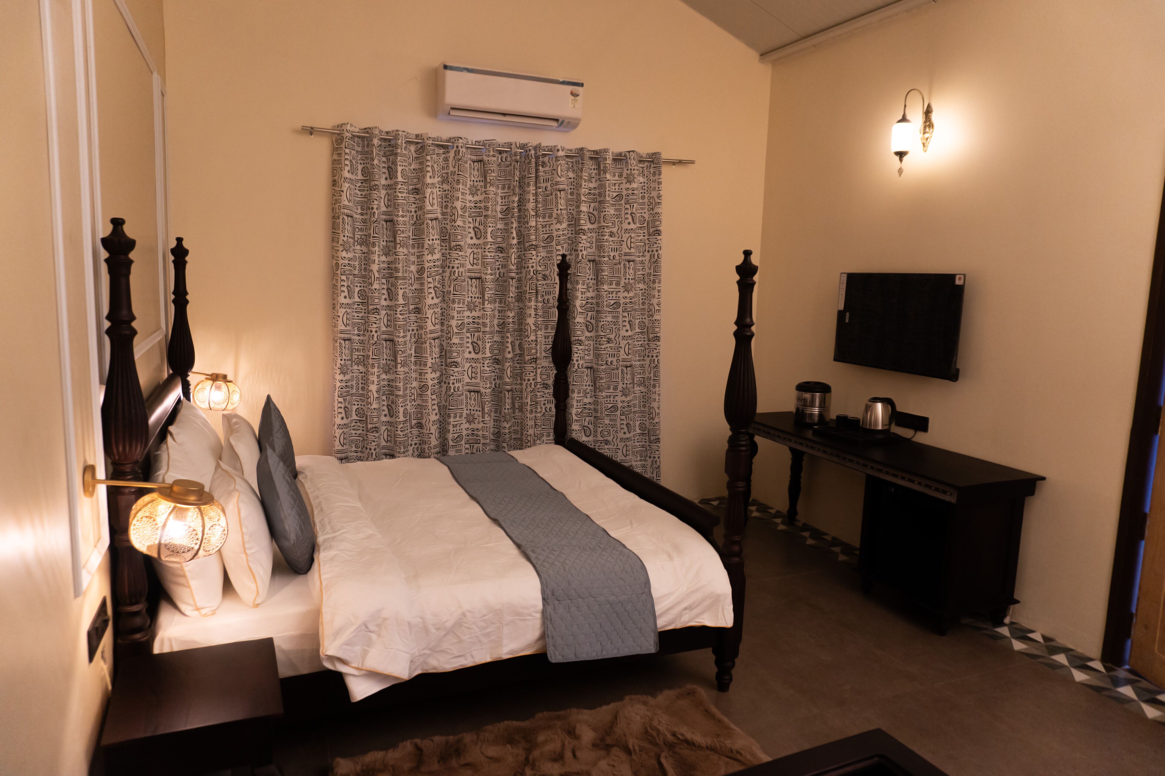 A bedroom with a bed, bedside table, lamp, air conditioning unit, television, and patterned curtains.