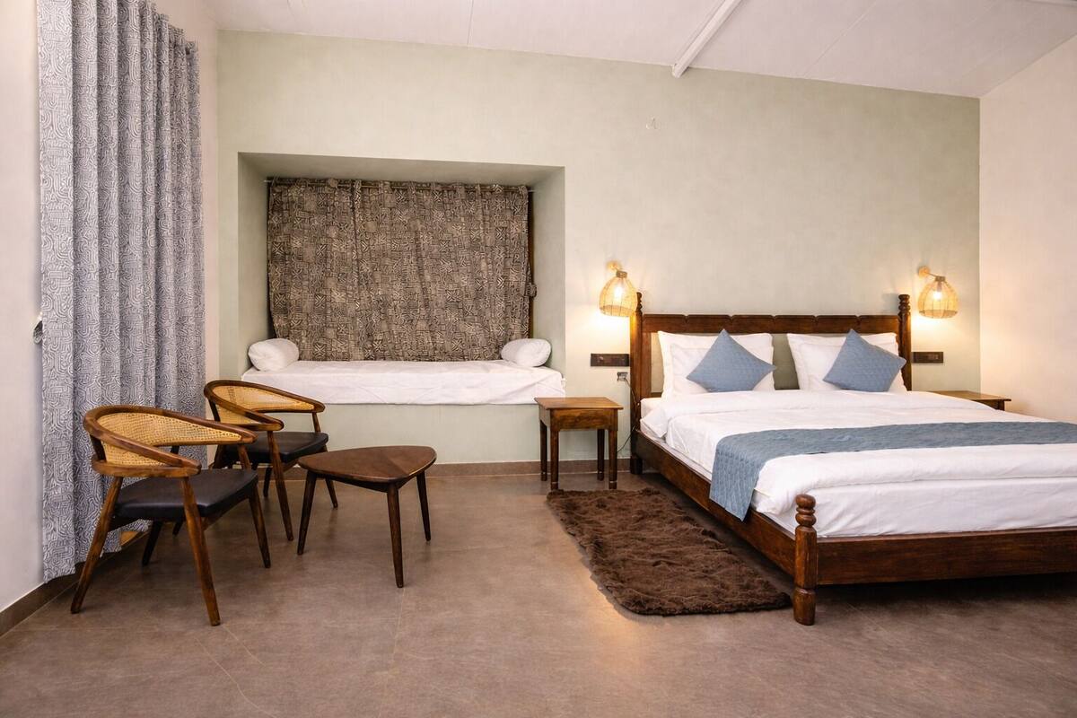 Une chambre à coucher comprenant un lit, deux tables de chevet, une chaise, une petite table, une fenêtre avec des rideaux à motifs et un luminaire fixé au mur.