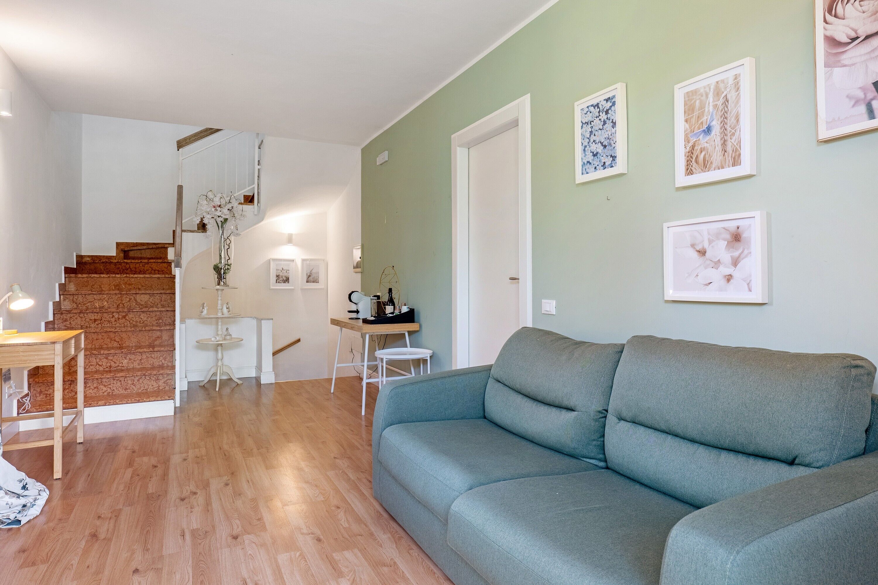 A living room with a grey sofa, wooden flooring, a staircase, and a small table with a lamp.