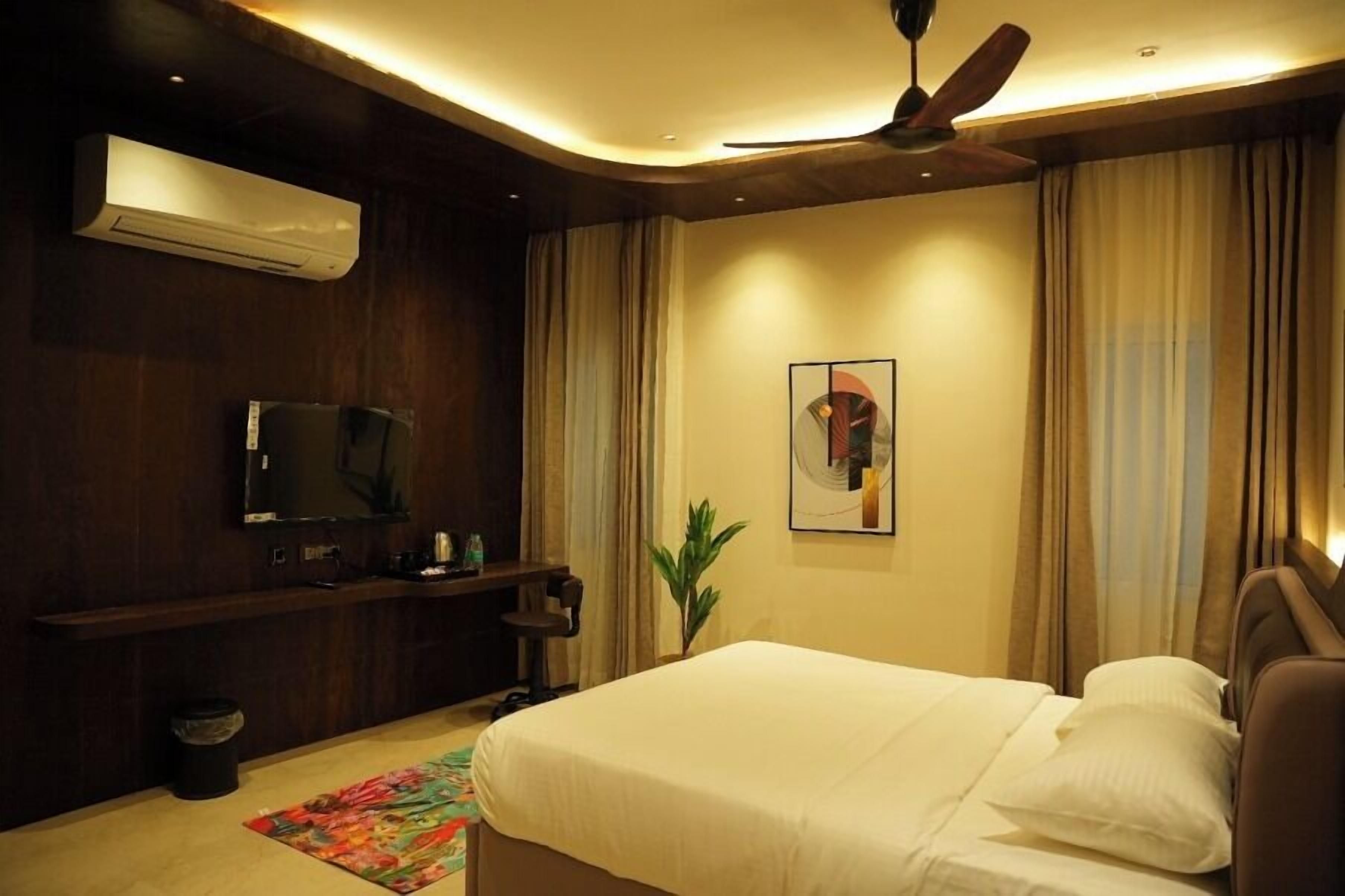 A hotel room with a bed, a ceiling fan, a TV, a plant, and a window with curtains.