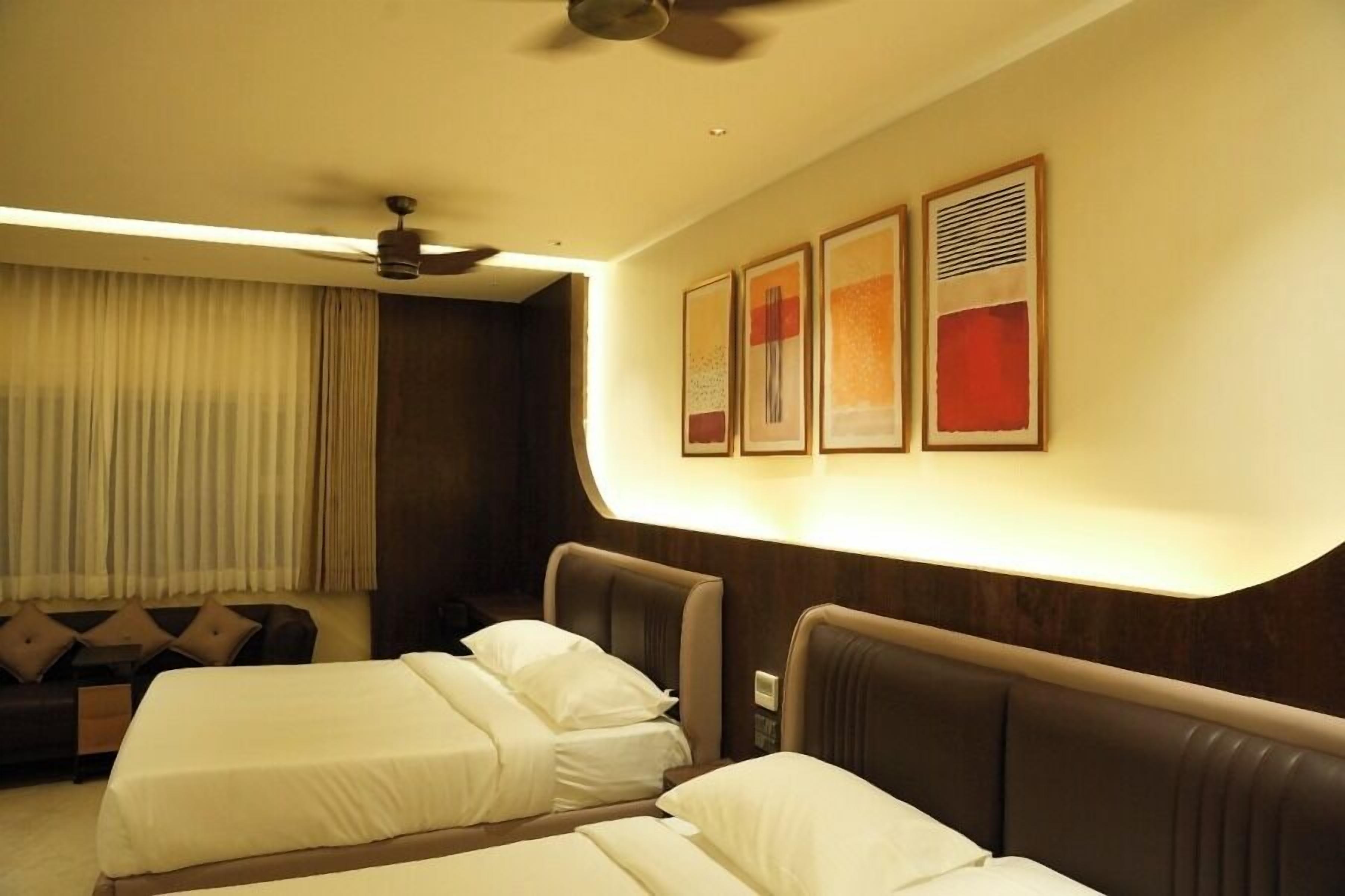 Habitación de hotel con dos camas, ventilador de techo, sofá y cuadros en la pared.