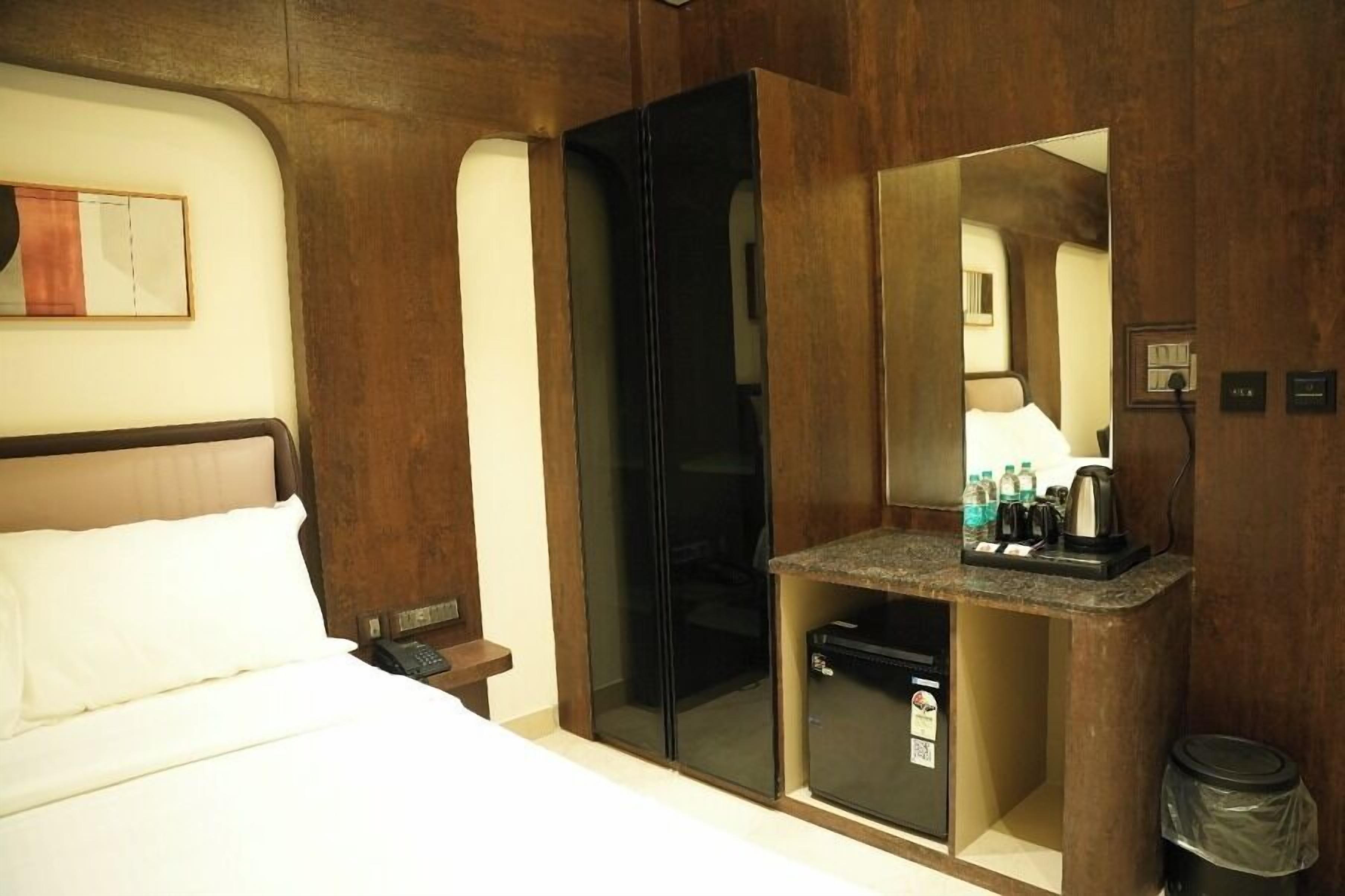 Habitación de hotel con cama, escritorio integrado con espejo, kitchenette pequeña y un bote de basura.
