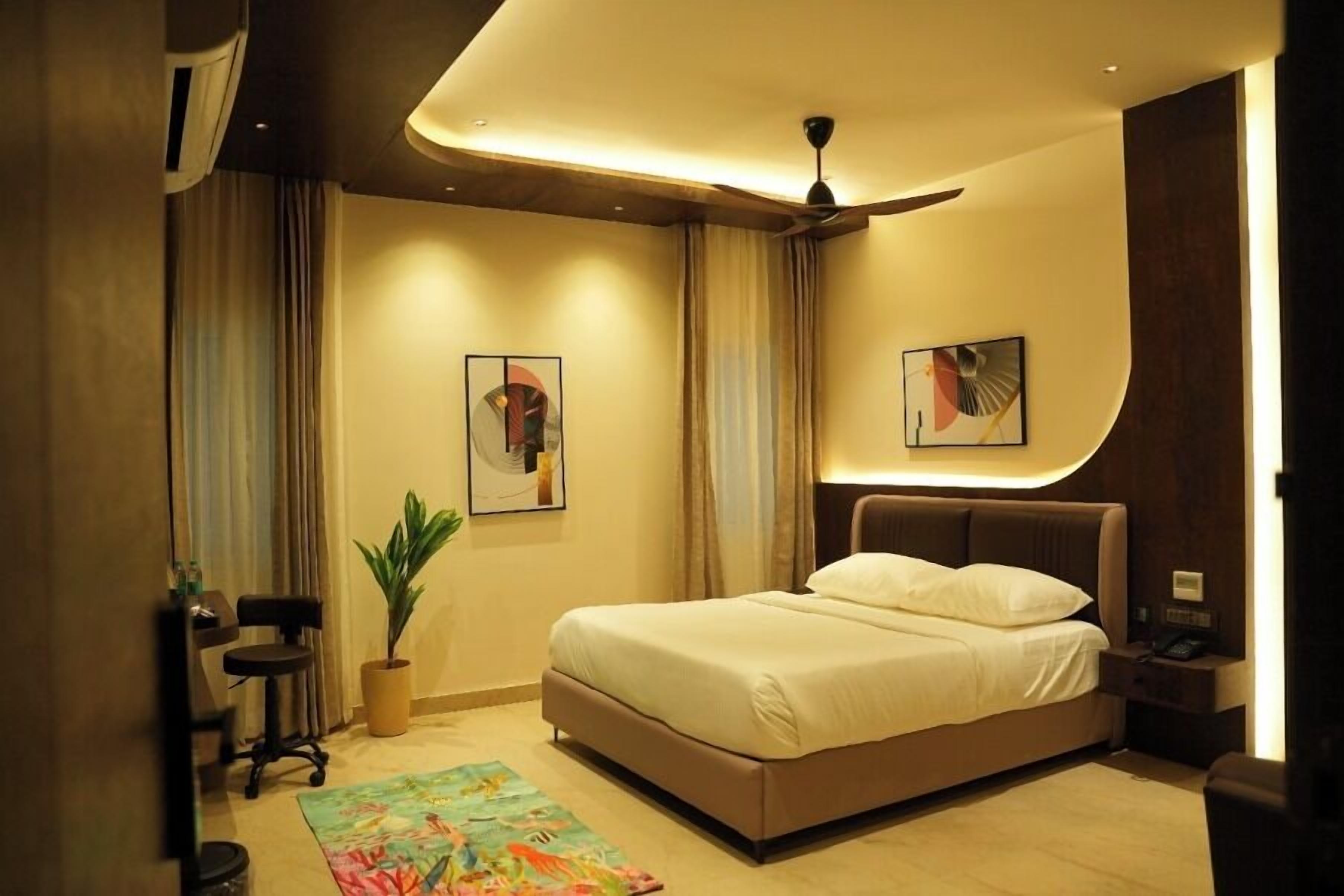 A hotel room with a bed, a ceiling fan, a desk with a chair, a potted plant, and artwork on the walls.
