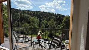 Ein Balkon mit Glasdoor, Gartenmöbeln und Blick auf einen bewaldeten Hügel.