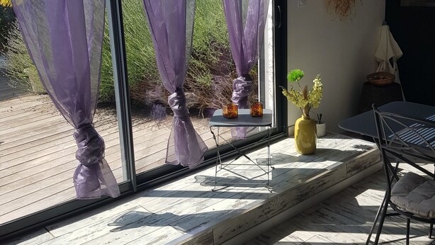 Una habitación con ventanas amplias, una mesa, sillas, un jarrón amarillo y una planta colgante.