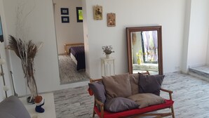 Una sala de estar con piso de madera, un sofá rojo, un sofá gris, una mesa de centro de madera, un espejo y una cama al fondo.