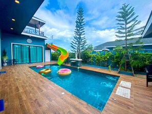 Una piscina con tobogán, rodeada de plantas tropicales y una terraza de madera.