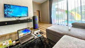 Ein modernes Hotelzimmer mit Flachbildfernseher, einem Laptop, einem Desktop-Computer, einer Couch und einem großen Fenster mit Blick auf einen Pool und Rutschen.