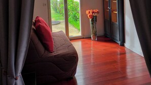 A room with a sliding glass door leading to a garden, a dark-colored sofa with a red pillow, a tall vase with flowers, and a dark wooden cabinet. - LA PETITE MAISON DE KERTHOMAS - CLASS 3* GÎTE (plumergat)