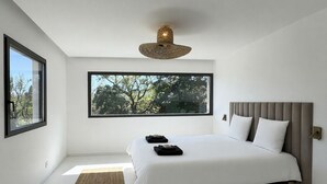 Ein modernes Schlafzimmer mit einem großen Bett, einem Fenster mit Blick auf Bäume, einer gewebten Deckenleuchte und einem kleinen Teppich.