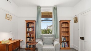 Ein Zimmer mit grauem Sessel, Holzbücherregalen, einem Schreibtisch mit Lampe und einem Fenster mit Vorhängen.