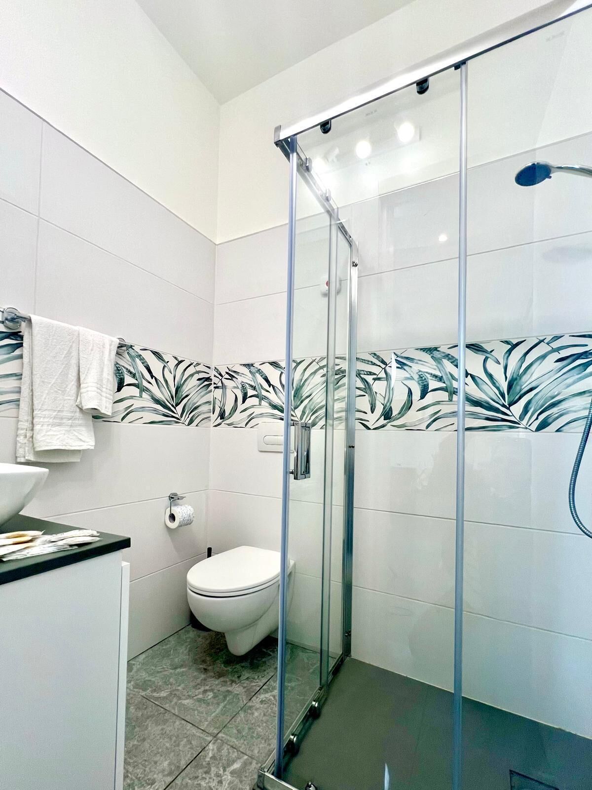 A bathroom with a glass shower enclosure, a toilet, a sink with a countertop, and a patterned wall.