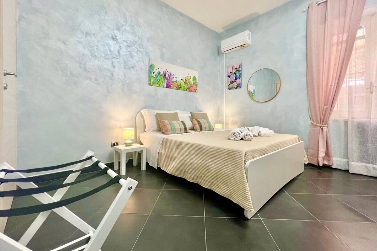 A bedroom with a bed, a nightstand, a lamp, an air conditioner, a mirror, and a window with curtains.