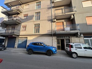 Ein mehrgeschossiges Gebäude mit Balkonen, ein blaues Auto davor und ein weißes Auto daneben.