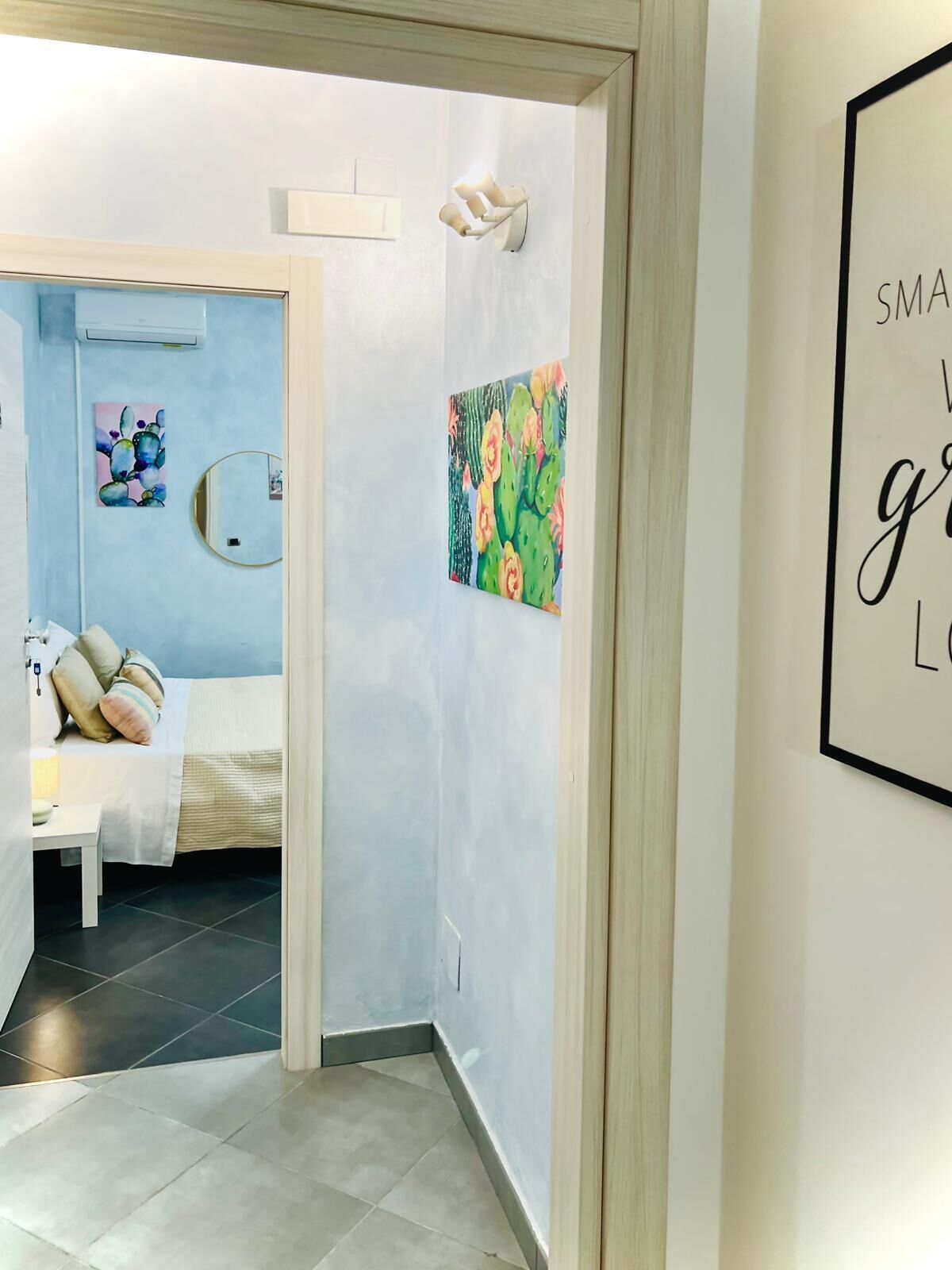 A hotel room with a bed, a bedside table, a wall-mounted mirror, and a painting of cacti.