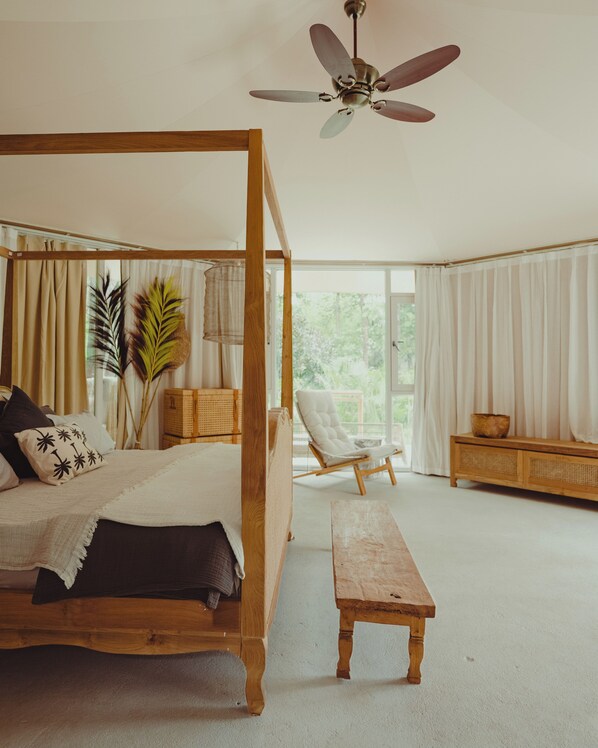 Una cama con dosel y tablero de madera, un banco de madera, un sillón acolchado y un ventilador de techo.