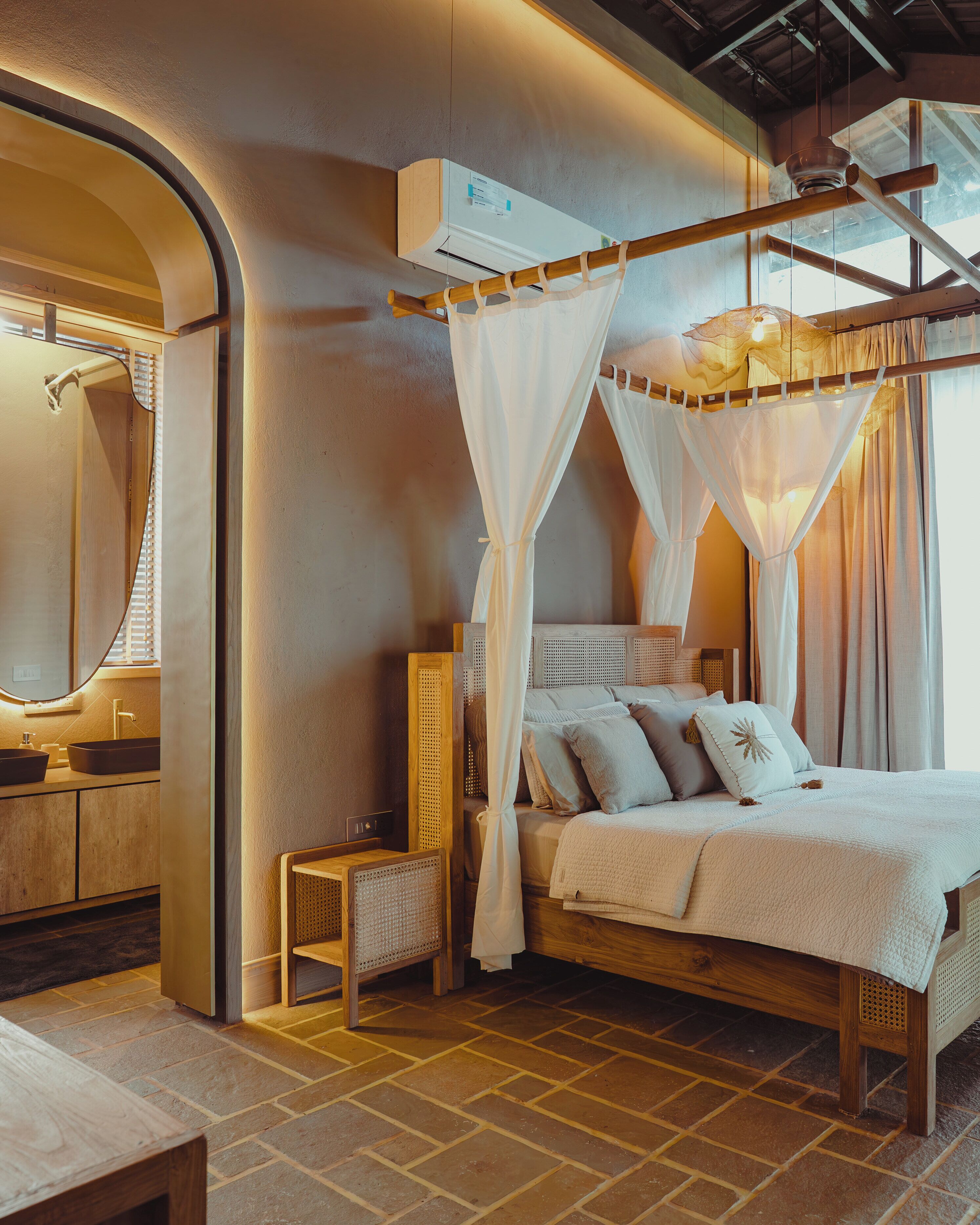 A bedroom with a canopy bed, a bedside table, a chair, a wooden floor, and an air conditioning unit.
