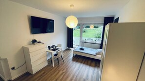 Ein gemütliches Zimmer mit Bett, Schreibtisch, Stuhl, Fernseher und einem Fenster mit Blick auf einen Garten.