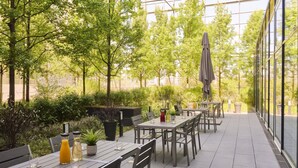 An outdoor patio with tables and chairs, surrounded by greenery and a glass wall. - Premier Inn München City West (Munich)