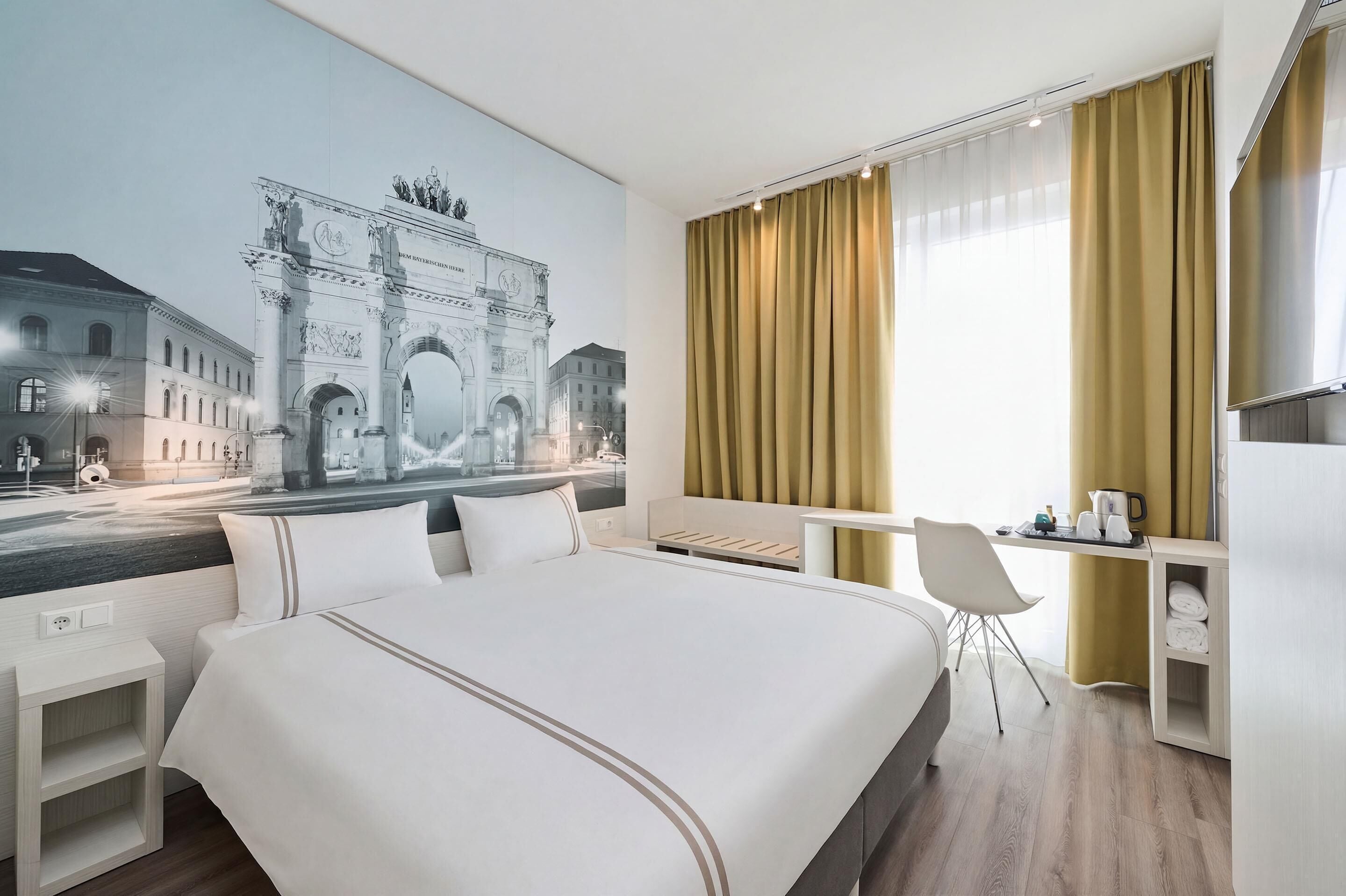 A hotel room with a large bed, a desk with a chair, a small table, and a wall-mounted TV.