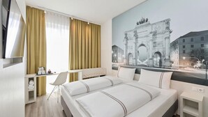 A hotel room with a bed, a desk, a chair, a TV, and a large window with curtains. - Premier Inn München City West (Munich)