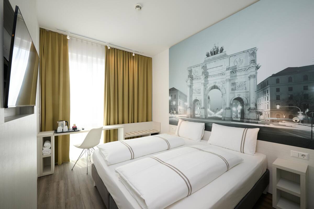 A hotel room with a bed, a desk, a chair, a TV, and a large mural of a historic arch.