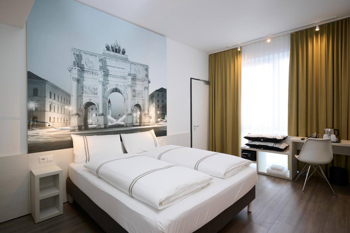 A hotel room with a large bed, a desk, a chair, a small bedside table, and a wall-mounted flat-screen TV.