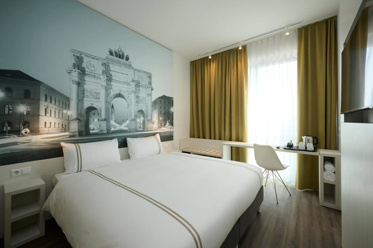 A hotel room with a large bed, a desk, a chair, a small table, and a large window with curtains.