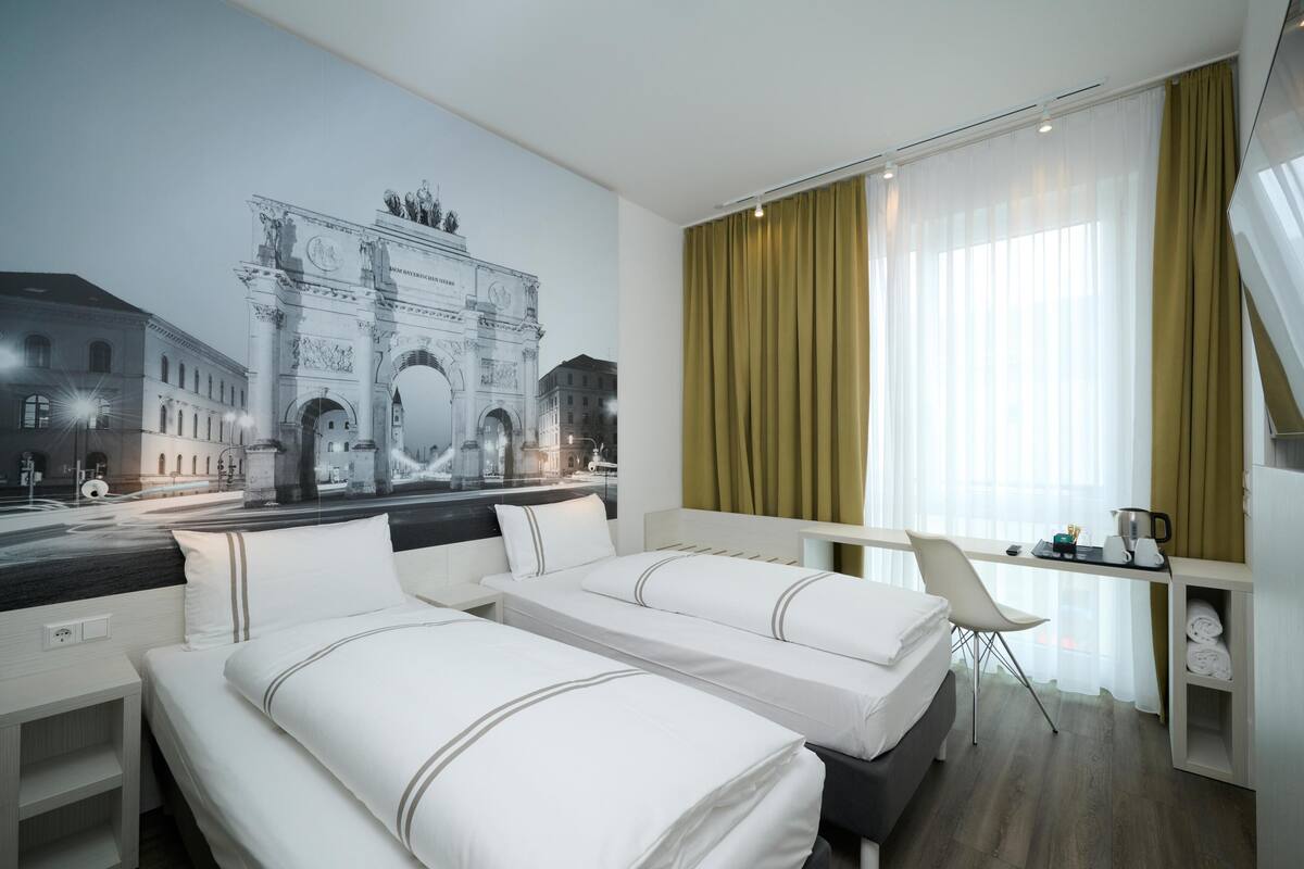 A hotel room with two beds, a desk, a chair, a window with curtains, and a mural of a historical building.