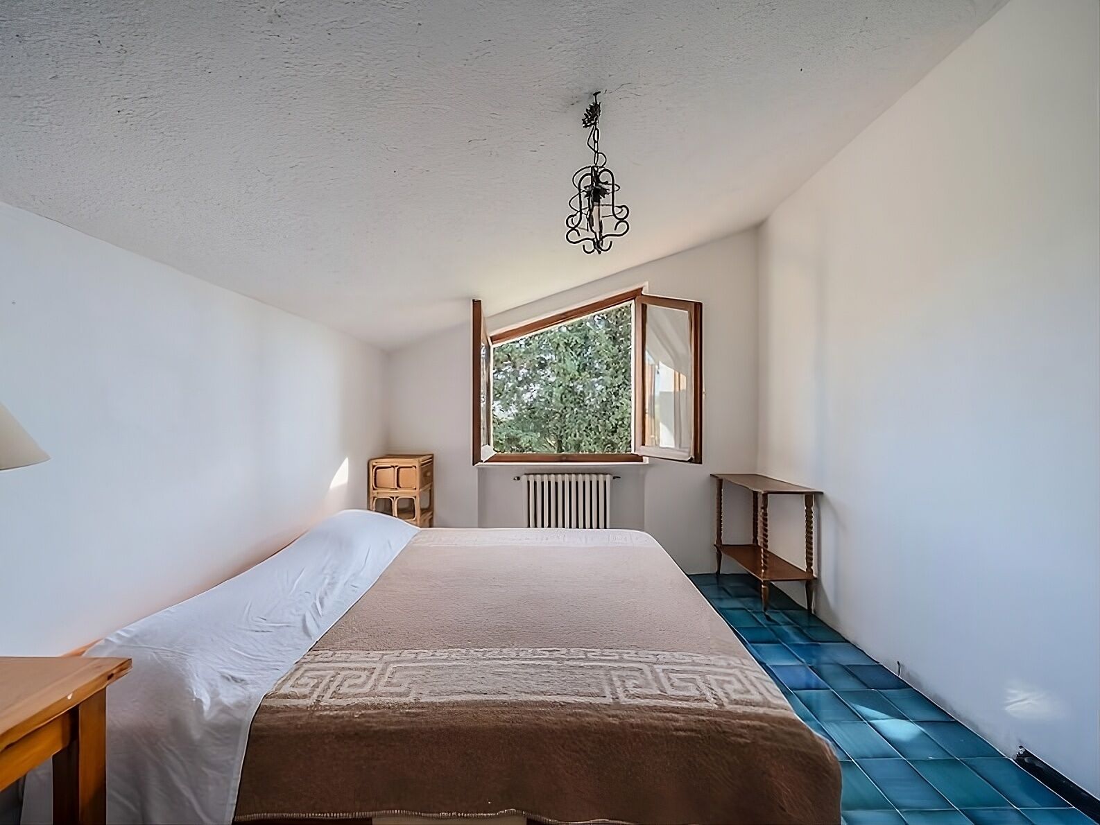 A bedroom with a bed, a window, a radiator, a wooden stool, and a small table.