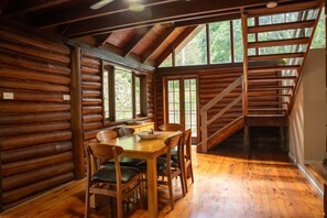 Interior de una cabaña de troncos con una mesa de comedor, sillas, una escalera y ventanas amplias.